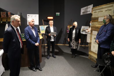 Grupa pięciu osób stoi wewnątrz pomieszczenia w otoczeniu obrazów i elementów związanych z tematyką kolejnictwa. Mężczyzna na środku trzyma mikrofon i wydaje się przemawiać, podczas gdy pozostali uczestnicy uroczystości słuchają. W tle widać plakaty, obrazy i drewniane skrzynie, które tworzą ekspozycję nawiązującą do historii kolei.