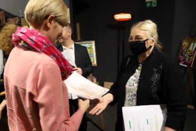 Dwie kobiety stoją naprzeciwko siebie i wręczają sobie dokumenty w towarzystwie innych osób. Jedna z kobiet, w różowym ubraniu, odebrała właśnie certyfikat czy dyplom. Na tle widać część ekspozycji z obrazami, świadczących o charakterze wydarzenia związanym z malarstwem i kolejnictwem.