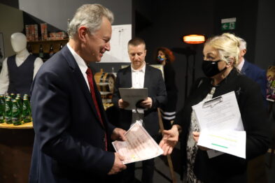 Mężczyzna i kobieta stoją naprzeciwko siebie, trzymając dokumenty i wpatrując się w nie; mężczyzna ma na sobie ciemny garnitur i krawat, a kobieta jasną bluzkę i ciemny żakiet. W tle widoczny jest mężczyzna w koszuli, który również przegląda dokumenty. Otoczenie zdaje się być wnętrzem z różnymi eksponatami, w tym odzieżą na manekinach.