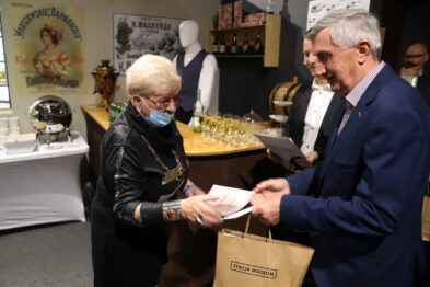 Starsza kobieta odbiera nagrodę z rąk mężczyzny, który trzyma dokumenty i torebkę z logiem. W tle eksponaty kolejowe i plakaty. Oboje są ubrani elegancko, a kobieta ma na twarzy maskę ochronną.