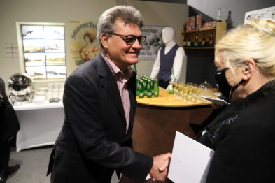 Mężczyzna w garniturze podaje rękę i uśmiecha się do kobiety, trzymającej kartkę w dłoni. W tle widać ekspozycję z manekinem w historycznym stroju kolejarza i stół z butelkami. Wydarzenie odbywa się w pomieszczeniu z wystawą tematyczną.