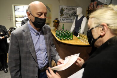 Osoba w maski ochronnej wręcza dokumenty mężczyźnie w szarej marynarce i koszuli bez krawata. W tle widoczne są manekiny ubrane w stroje z epoki, stoły z butelkami oraz plakaty kolei. Scena rozgrywa się w pomieszczeniu z wystawą tematyczną.