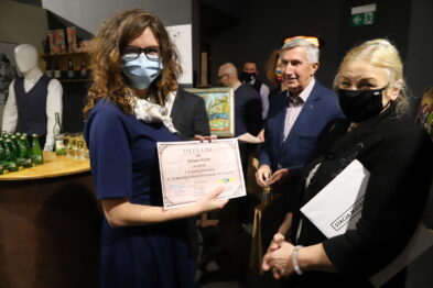 Trzy osoby stoją obok siebie, dwie kobiety i mężczyzna w środku, w przestrzeni wystawienniczej. Kobieta z lewej przyjmuje dyplom, ma na sobie niebieską sukienkę i maseczkę ochronną. Na drugim planie widoczne są elementy wystawy związane z koleją, w tym manekin w historycznej mundurze.