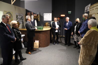 Grupa osób stoi w pomieszczeniu, w którym dominuje ciemne tło i sztuczne oświetlenie. W centrum grupy znajduje się mężczyzna w jasnej koszuli, który coś prezentuje lub opowiada, a pozostali uczestnicy wydają się uważnie słuchać. W tle widać wystawione modele pociągów oraz plansze informacyjne.