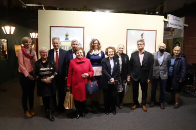 Grupa osób stoi uśmiechnięta do kamery na tle wystawy z okazji 160. rocznicy Kolei Petersbursko-Warszawskiej. W centralnej części zdjęcia kobieta w czerwonym płaszczu trzyma drobny przedmiot. W tle widać plansze informacyjne i fotografie związane z historią kolei.