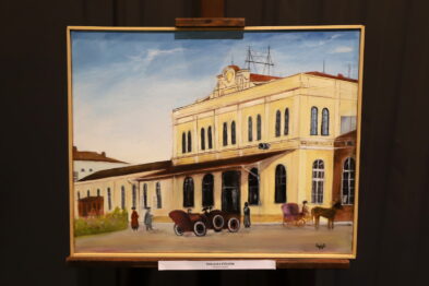 Obraz przedstawia budynek stacji kolejowej z epoki, z dwuwózkiem konnym i poczekalnią na pierwszym planie. Dzieło jest malarskie, kolorowe, zawieszono je na sztaludze. Szczegóły architektoniczne i elementy krajobrazu są wyraźnie zaznaczone.
