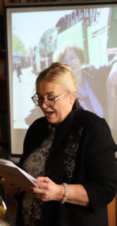 Kobieta stoi przed mikrofonem i czyta z kartki; tło za nią tworzy duży ekran, na którym wyświetlane są zdjęcia związane z kolejnictwem. Jest ubrana w ciemny garnitur i stoi w pomieszczeniu z półkami pełnymi książek. Oświetlenie w pomieszczeniu jest ciepłe, tworząc przytulną atmosferę.