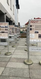 Widoczne są metalowe tablice ekspozycyjne umieszczone na chodniku przed budynkiem. Tablice zawierają informacje i fotografie związane z historią kolei. Na pierwszym planie znajduje się tablica z napisem „Stacja Muzeum".