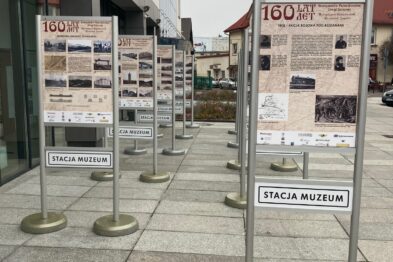 Widoczne są metalowe tablice ekspozycyjne umieszczone na chodniku przed budynkiem. Tablice zawierają informacje i fotografie związane z historią kolei. Na pierwszym planie znajduje się tablica z napisem „Stacja Muzeum