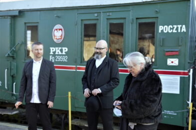 Trzy osoby stoją obok siebie przed zabytkowym zielonym wagonem kolejowym z napisem 