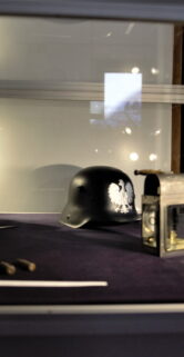 W witrynie ekspozycyjnej znajdują się przedmioty związane z koleją, w tym stara latarnia kolejowa, hełm i inne akcesoria. Na pierwszym planie witryny umieszczono model lokomotywy oraz białe rękawiczki. Część eksponatów jest oświetlona, co pozwala na lepsze ich wyeksponowanie na tle ciemnofioletowego wnętrza witryny.