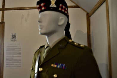 Manekin ubrany w wojskowy mundur z epoki, z czapką na głowie, prezentuje uniform powstańczy. Na mundurze widoczne są odznaczenia i insygnia. W tle częściowo widoczna jest informacyjna tablica wystawy.