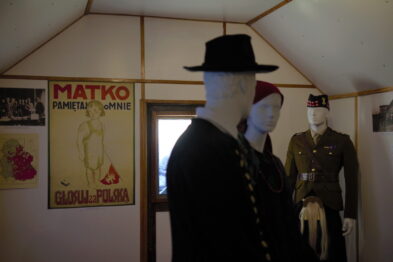 Manekiny ubrane są w stroje z epoki: mężczyzna w ciemnym garniturze z kapeluszem, a drugi w mundurze wojskowym z nakryciem głowy. Ściany ozdobione są plakatami, w tym jednym z napisem 