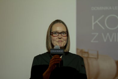 Kobieta stoi na scenie trzymając mikrofon i prezentuje się publiczności. W tle znajduje się baner promujący książkę zatytułowaną 