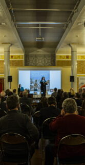 W eleganckiej, przestronnej sali zgromadziła się publiczność na krzesłach skierowana ku prelegentom; scenę zdobią złote zdobienia i duży ekran prezentacyjny. Dwóch mówców stoi przy mównicy, jeden przemawia, drugi słucha; obaj są ubrani w ciemne garnitury. Ściany ozdobione są klasycznymi detalami architektonicznymi, a oświetlenie dodaje uroczystego charakteru wydarzeniu.