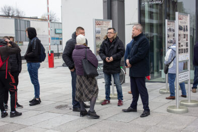Grupa ludzi gromadzi się przed panelem informacyjnym na zewnątrz. Osoby są ubrane w zimowe odzieży i wydają się skupione na rozmowach. Większość zwraca uwagę na wystawę, a w tle widać nowoczesny budynek.