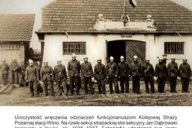 Grupa mężczyzn w mundurach stoi przed niewielkim budynkiem; wszyscy są ustawieni w dwóch rzędach, a niektórzy z nich trzymają kapelusze w rękach. Mężczyźni noszą odznaki, a ich ubiory wskazują na przynależność do służb kolejarza lub straży. Jeden z mężczyzn siedzi z lewej strony obok szyldu z napisem stacji.