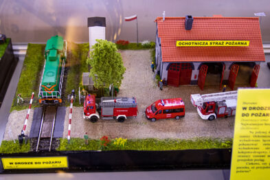 Miniaturka przedstawia makietę kolejową z zielonym lokomotywą, przecinającą przejazd kolejowe z zapory. Obok torów znajduje się budynek oznaczony jako straż pożarna, przed którym zaparkowane są czerwone pojazdy strażackie. Otoczenie dopełniają drzewa i równo przycięta trawa, nadając scenie realistyczny wygląd.