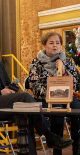 Dwoje osób siedzi na krzesłach przed choinką, kobieta trzyma książkę i coś opowiada, a mężczyzna obok uważnie jej słucha. W tle widać okno z łukowatym zwieńczeniem i ciepłe oświetlenie, które nadaje pomieszczeniu przytulną atmosferę. Na pierwszym planie widać egzemplarz książki wystawiony na niskim stołku.