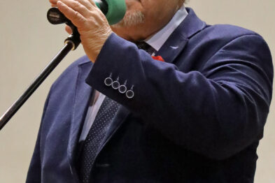 Starszy mężczyzna ubrany w ciemny garnitur śpiewa do mikrofonu trzymanego w prawej ręce. Występuje na scenie przed mikrofonem, jego mimika twarzy wskazuje na emocjonalne zaangażowanie w śpiew. Na jego lewej piersi widoczne są dekoracje lub odznaczenia.