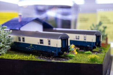 Model kolejowy przedstawia dwa zielone wagony pasażerskie z białymi pasami i czarnymi dachami umieszczone są na makiecie z trawą i krzewami. Na tle widać makietę budynku oraz postać ludzką w żółtej koszulce. Całość jest starannie wykonaną imitacją sceny kolejowej w skali miniatury.