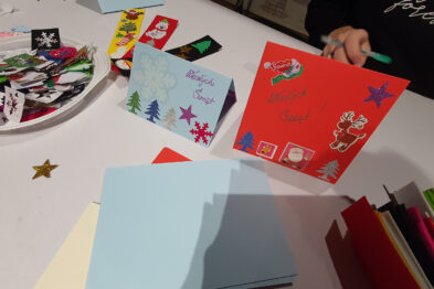 Na stole rozłożone są kolorowe kartki i wycinanki, z których uczestnicy tworzą kartki świąteczne. Widać rękę osoby trzymającej jedną z kartek, ozdobioną rysunkami i naklejkami. Obok leżą inne przykłady kartek i wycinanek, gotowe do dalszej pracy kreatywnej.