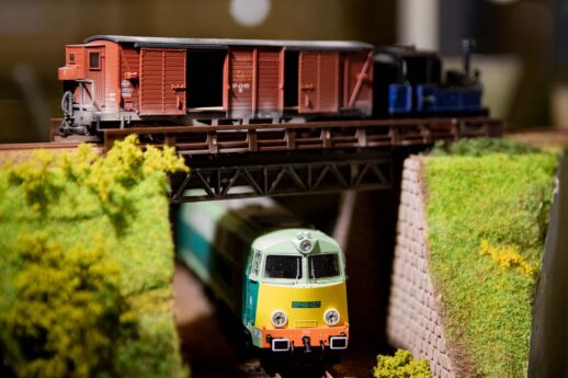 Widoczne są dwa modele pociągów znajdujące się na makiecie kolejowej. Górna część obrazu przedstawia czerwony lokomotyw towarowy przejeżdżający przez stalowy most, a w dolnej części zdjęcia widać model pociągu pasażerskiego, pomalowanego w jasnozielone i kremowe kolory, który wydaje się przejeżdżać pod mostem. Całość otaczają elementy krajobrazu, takie jak modele drzew, trawa oraz makietowe nasypy.
