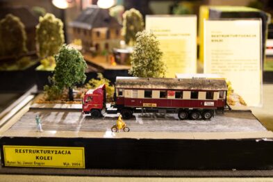 Miniatura pociągu z wagonami stoi na podstawie obok miniaturowych drzew i figurki ludzi. Obok torów zaparkowany jest czerwony model samochodu ciężarowego z naczepą. Tło stanowi informacyjna kartka z tekstem i kolejne modele kolejowe.