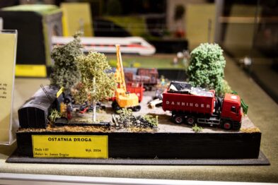 Model kolejki przedstawiający scenę na tle krajobrazu z drzewami i postaciami ludzkimi. Czerwona lokomotywa w towarzystwie kilku wagonów jest na torach, a obok widać małą koparkę i załadunek. Na pierwszym planie umieszczono tabliczkę z napisem 