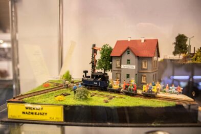 Model kolejki parowej z lokomotywą i dwoma wagonami jest przedstawiony na makiecie zielonego terenu z domkiem, drzewkiem i postaciami ludzkimi. Makietę charakteryzują detale takie jak nasadzenia, figurki ludzi w różnych kolorach oraz tory kolejowe. W tle na etykiecie widnieje napis 