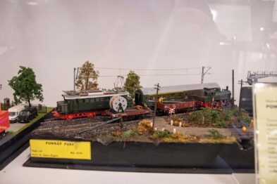 Model kolejowy jest prezentowany na gablotce wystawowej, przedstawiający zieloną lokomotywę i czerwony wagon na torach. Dookoła torów umieszczono miniaturowe elementy scenerii – drzewa, budynki i postaci. Model umieszczony jest na podstawie z etykietką informacyjną.