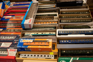 Modele pociągów leżą obok siebie na powierzchni, zapakowane w oryginalne opakowania o różnych kolorach i rozmiarach. Każde opakowanie posiada nadruk z nazwą oraz grafiką reprezentującą zawartość. Modele te są ułożone w rzędy tworząc kolorowy mozaikowy efekt.