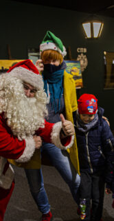 Osoba przebrana za Świętego Mikołaja pochyla się, aby porozmawiać z grupą dzieci, które patrzą na nią z zainteresowaniem i uśmiechem. W tle widoczne są kolorowe ilustracje oraz historyczne zdjęcia związane z kolejnictwem. Dzieci mają na sobie ciepłe zimowe ubrania; jedno z nich nosi świąteczną czapkę, a inne są ubrane w kurtki i czapki chroniące przed chłodem.