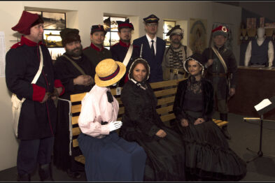 Grupa osób ubrana w historyczne kostiumy stoi i siedzi w środku pomieszczenia; kilka kobiet w sukniach z epoki i mężczyźni w mundurach, jeden z nich w błękitnej kurtce z czerwonymi wykończeniami i czapką z daszkiem. W tle widoczne są manekiny także ubrane w stroje historyczne, a cała scena jest oświetlona sztucznym światłem. Atmosfera zdjęcia wydaje się uroczysta i poważna.