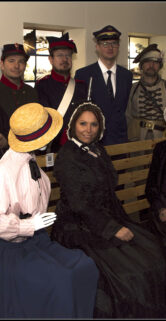 Grupa osób ubranych w historyczne stroje stoi i siedzi wewnątrz pomieszczenia podczas rekonstrukcji historycznej. Wśród strojów dominują mundury wojskowe i eleganckie suknie damskie z okresu XIX wieku, a także tradycyjne nakrycia głowy takie jak kepi i kapelusze. Tło stanowi wystawa z manekinami prezentującymi inne stroje epoki, a przestrzeń jest oświetlona sztucznym światłem.