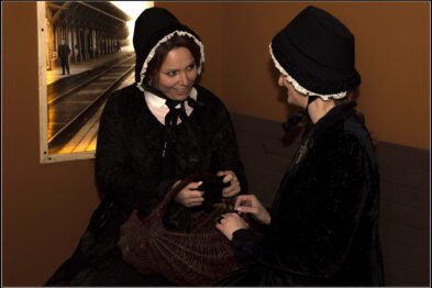 Dwie kobiety w historycznych strojach siedzą naprzeciwko siebie i są zaangażowane w rozmowę. Jedna z nich trzyma przedmiot, który może być częścią stroju lub rekwizytem. W tle widoczne jest zdjęcie przedstawiające stację kolejową z tamtej epoki, co wzmacnia historyczny charakter sceny.