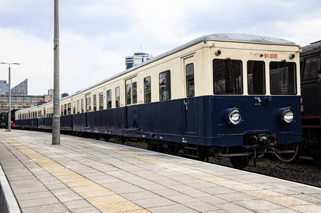 The electric traction unit E91, marked with the number EW51, is standing on the platform next to another train set. The unit has a dark blue livery with white elements and a cream-colored upper part. The set consists of several cars, and its side doors and windows indicate an older model of the vehicle.