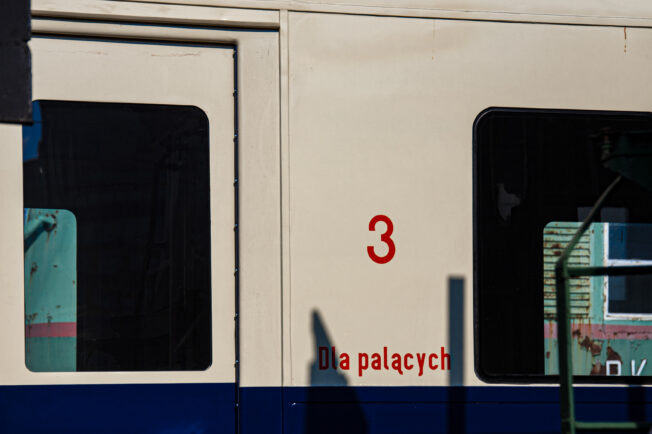 The visible part of a blue-beige railway carriage with the sign 'for smokers' just below the red number '3'. The vehicle's doors are closed, and through the window, reflections and indeterminate structures can be seen. The construction of the vehicle indicates an older model, consistent with the historical context of suburban transport.