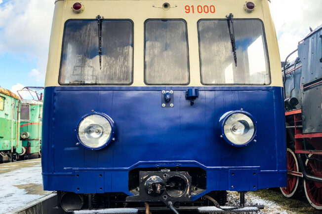 Elektryczny zespół trakcyjny E91 o oznaczeniu 91 006 stoi na torach. Pojazd ma niebiesko-białe malowanie oraz okrągłe reflektory z przodu. Zespół jest otoczony innym taborom kolejowym i znajduje się pod gołym niebem.