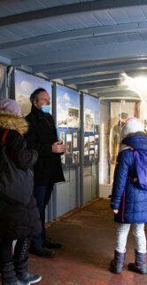Grupa osób zwiedza wnętrze wyłożone zdjęciami i plakatami tematycznymi związanymi z kolejnictwem. Ludzie, ubrani w zimowe kurtki, stoją i przemieszczają się po pomieszczeniu, przyglądając się eksponatom. Ściany i panele wystawiennicze zachowują kolorystykę związaną z tematyką kolejową.