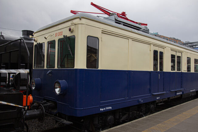 Elektryczny zespół trakcyjny E91 oznaczony numerem E1 stoi na torach kolejowych. Pojazd jest pomalowany na niebiesko z kremowym pasem na górnej części. Zespół wyposażony jest w pantograf i posiada okrągłe reflektory.