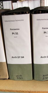 Widoczne są segregatory ustawione na półce, z etykietami wskazującymi na przynależność do różnych serii dokumentów technicznych lokomotyw oznaczonych jak PT31 czy arch. OT 84. Każdy segregator ma na grzbiecie napisy oraz unikatowy numer, zapewne służący klasyfikacji i identyfikacji. Segregatory mają jednolite kolory i są ułożone w porządku obok siebie.