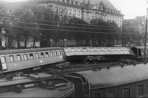 Na fotografii czarno-białej widoczne są wykolejone wagony kolejowe o różnych konstrukcjach, które leżą na boku między innymi torami. Przy jednym z torów stoją nienaruszone wagony pasażerskie. W tle majaczy budynek, który przypomina stację kolejową lub inny duży obiekt architektoniczny.