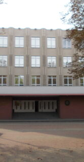 Na pierwszym planie znajduje się brukowana droga prowadząca do wejścia w wielopiętrowym budynku z cegły. Wokół drogi widać wyłożone płyty chodnikowe oraz kilka niskich drzew i latarni ulicznych. Budynek o symetrycznie rozmieszczonych oknach posiada czerwoną fasadę na parterze, a wyższe kondygnacje pokryte są jasnym tynkiem.