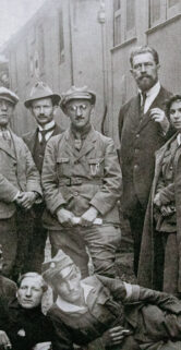 Grupa ludzi w historycznych strojach pozujących do zdjęcia; wśród nich widoczni są mężczyźni w mundurach wojskowych i kobiety w białych strojach z zasłonami na głowie. Za postaciami widać wagon kolejowy ze słabo widocznymi napisami. Osoby na zdjęciu stoją i siedzą w różnych pozach, tworząc scenę przypominającą rodzinną fotografię z dawnych lat.