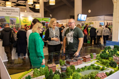 Trzy osoby rozmawiają przy makiecie kolejowej z detalami takimi jak budynki, drzewa i torowisko. Model przyciąga uwagę odwiedzających stoiska na targach, zainteresowanych wystawą. W tle dostrzegamy inne ekspozycje targowe i grupę zwiedzających.
