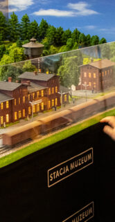 Dziecko stoi przed makietą kolejową prezentującą zabudowania stacyjne, obserwując szczegóły modelu. Makietę chroni przezroczysta szyba, a na przedniej krawędzi znajduje się opis "STACJA MUZEUM". W tle widać różne modele terenów i budynków kolejowych w miniaturowej skali, starannie wykonane i kolorowe.