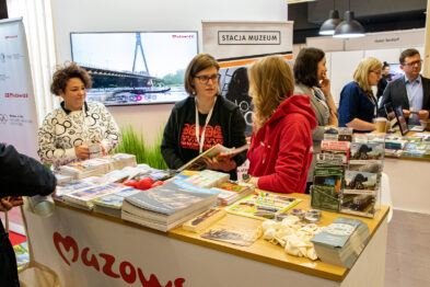 Osoby stojące i siedzące przy stoisku targowym rozmawiają i przeglądają różnorodne materiały, takie jak broszury czy książki. Stoisko jest wyposażone w jasną osłonę z napisami oraz posiada na blacie ułożone pamiątki i wydawnictwa. Wśród eksponatów widać także makietę mostu, co nawiązuje do tematu kolejnictwa.