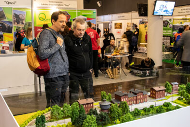 Dwóch mężczyzn zwiedza makietę kolejową, którą otaczają bujne, zminiaturyzowane drzewa i modele budynków. Jeden z nich, odziany w czerwoną kurtkę, z zainteresowaniem wskazuje palcem, prowadząc rozmowę z drugim mężczyzną. W tle dostrzegać można inne ekspozycje targowe i odwiedzających.