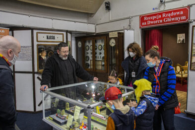 Grupa osób ogląda eksponaty umieszczone w szklanej witrynie w pomieszczeniu z zegarami i tabliczkami związane z tematyką kolejową. W środku witryny znajdują się modele pociągów i związane z nimi obiekty miniaturkowe. Ubrany na czarno mężczyzna gestykuluje i wskazuje na przedmioty w witrynie, objaśniając szczegóły grupie słuchaczy, wśród której jest także młody chłopiec.